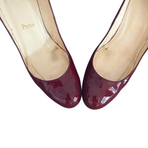 Par de zapatos de tacón de charol burdeos. Los zapatos tienen una puntera redondeada y un acabado brillante. El interior del zapato es de color beige claro con la palabra "París" impresa en letras doradas.