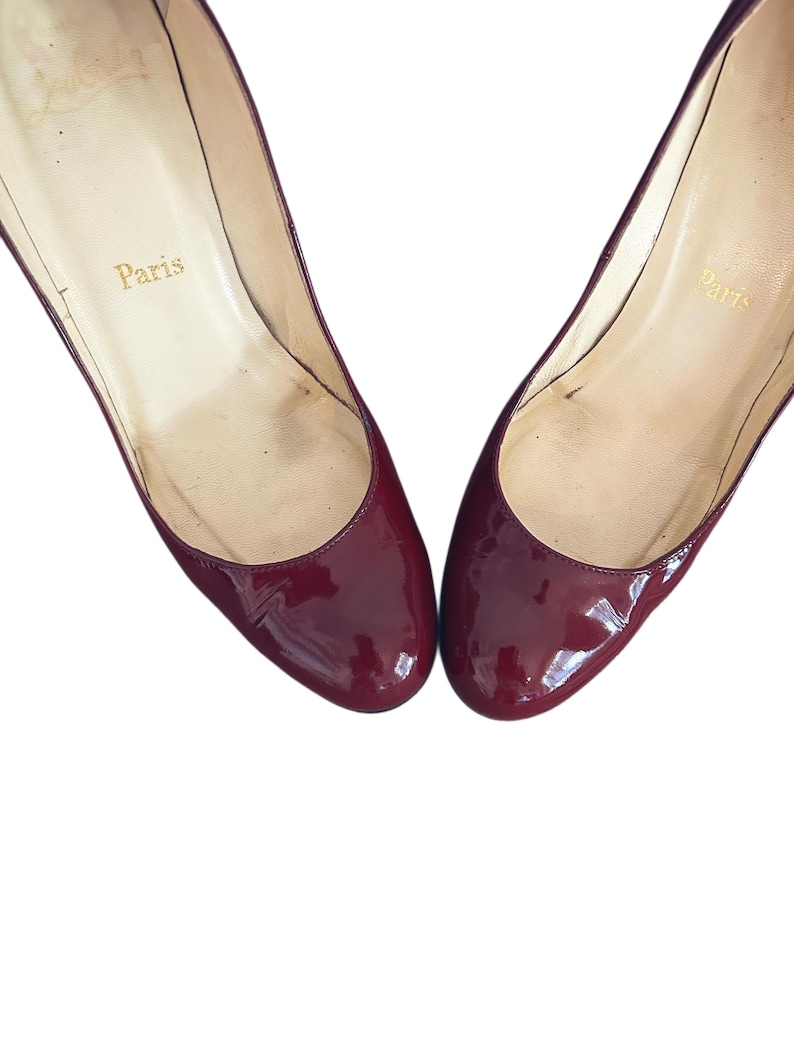 Par de zapatos de tacón de charol burdeos. Los zapatos tienen una puntera redondeada y un acabado brillante. El interior del zapato es de color beige claro con la palabra "París" impresa en letras doradas.
