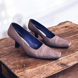 May include: A pair of mauve-colored, low-heeled pumps with a square toe and small black heels. The shoes have a dotted pattern and a blue interior. The shoes are displayed on a wooden surface.