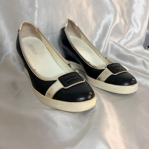 Puede incluir: Un par de zapatillas de cuña Guess en blanco y negro. Los zapatos tienen una parte superior de cuero negro con suela y ribete blancos. El logotipo de Guess es visible en la lengüeta y en un pequeño cuadrado negro en la parte superior del zapato.