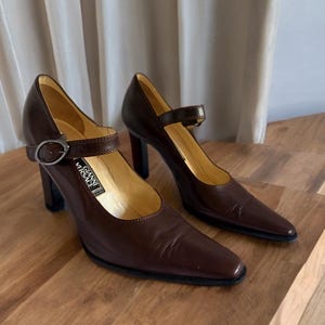 以下が含まれることがあります： 茶色の革製ハイヒールパンプス。上部にストラップとバックル留めが付いています。靴はポインテッドトゥとブロックヒールです。「Gianni Versace」のブランド名がインソールに見えます。