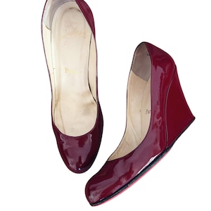 May include: Pair of glossy, burgundy wedge heels. The shoes have a rounded toe and a wedge heel. The soles are red. The word "Paris" is visible inside the shoe.