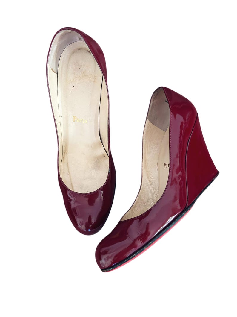 May include: Pair of glossy, burgundy wedge heels. The shoes have a rounded toe and a wedge heel. The soles are red. The word "Paris" is visible inside the shoe.