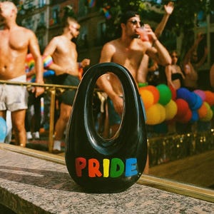 Puede incluir: Un objeto decorativo negro en forma de lágrima con una gran abertura ovalada. La palabra "PRIDE" se muestra en colores del arcoíris en la parte inferior. El fondo muestra un desfile con gente y globos coloridos.