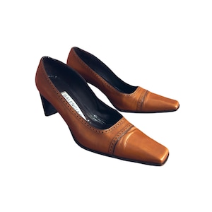 Puede incluir: Un par de zapatos de tacón de cuero marrón con punta cuadrada y tacón de bloque. Los zapatos tienen costuras decorativas en la parte superior y la marca "BALENCIAGA" es visible en la plantilla.