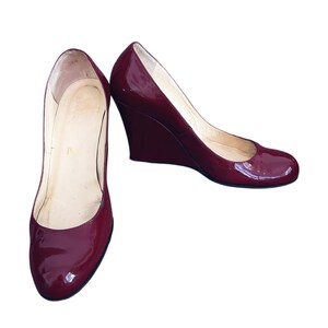 May include: A pair of burgundy patent leather wedge heels. The shoes have a rounded toe and a wedge heel. The interior of the shoe is a light beige colour. The shoes are set against a white background.
