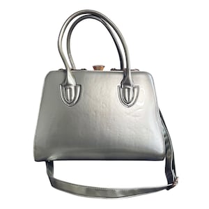Puede incluir: Un bolso de mano metálico plateado con asa superior y correa de hombro desmontable. El bolso tiene un cierre dorado.