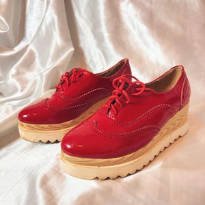 Peut inclure: Une paire de chaussures à plateforme en cuir verni rouge avec une semelle aspect bois. Les chaussures ont un design à lacets et des perforations décoratives. La semelle plateforme mesure environ 5 cm de haut.