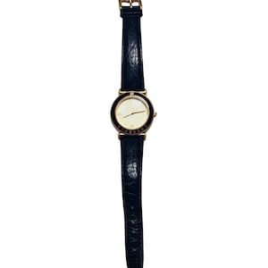 Puede incluir: Un reloj de pulsera dorado con una correa de cuero negro. La esfera del reloj es blanca con agujas doradas y un bisel negro. La marca "GIVENCHY" está impresa en la esfera.