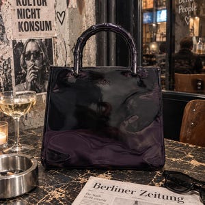 May include: A black patent leather handbag with a glossy finish and a dark bamboo-style handle. The front of the bag features the embossed text Gucci made in Italy. The bag has a structured, square silhouette with clean lines and a polished appearance.