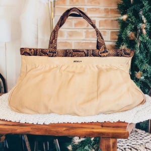 Puede incluir: Un bolso de mano beige con ribetes y asas con estampado de piel de serpiente marrón. El bolso tiene un cuerpo fruncido de textura suave con el nombre de la marca FURLA impreso en letras negras pequeñas en el centro, bajo el borde estampado.