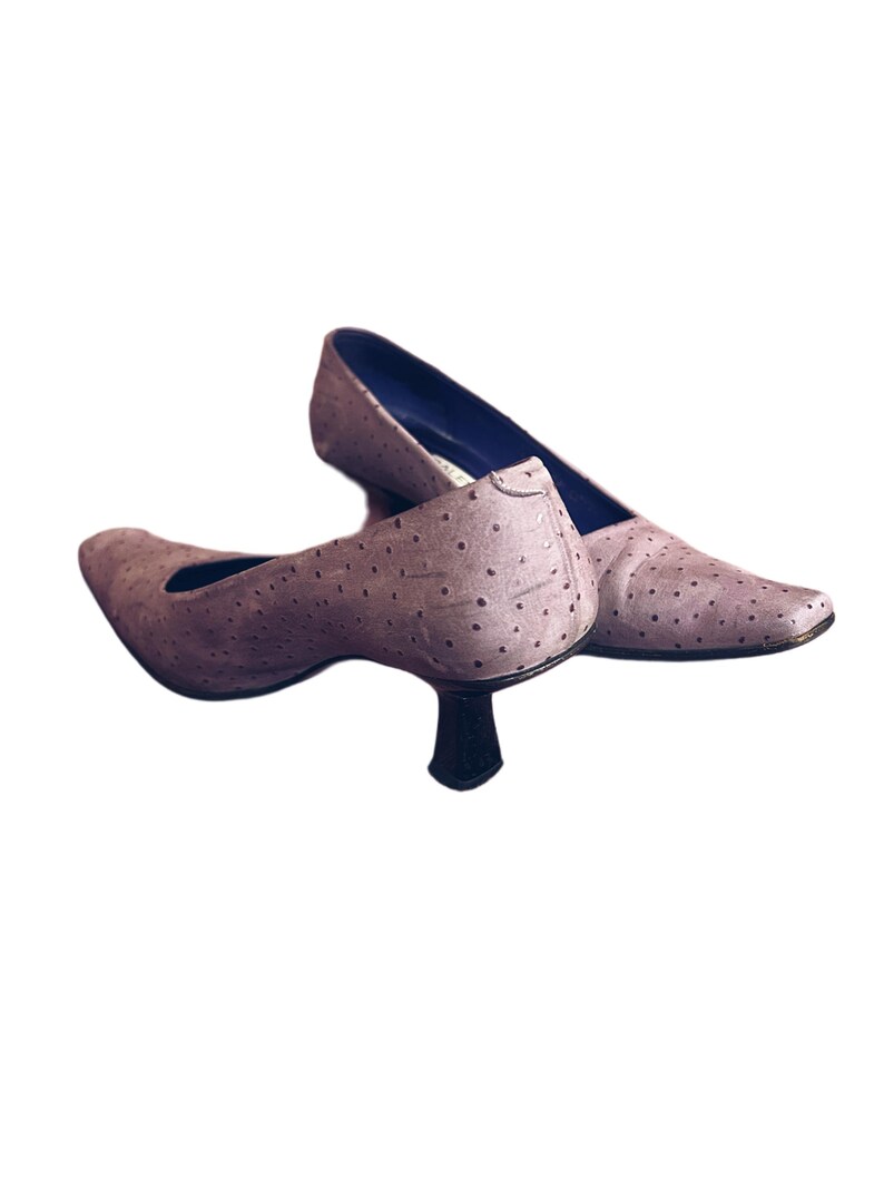 May include: A pair of light purple pumps with a pointed toe and a small, black heel. The shoes have a textured surface with small, evenly spaced dots. The interior of the shoe is a dark blue color.