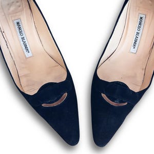 Puede incluir: Un par de zapatos de tacón Manolo Blahnik de ante negro. Los zapatos tienen una punta afilada y un pequeño detalle decorativo en la pala. El interior del zapato es de color beige claro y el nombre de la marca está impreso en la plantilla.