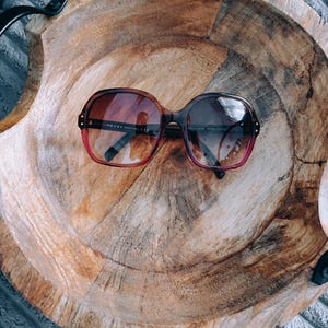 Puede incluir: Gafas de sol de gran tamaño con montura degradada de marrón a rosa y lentes oscuras. La patilla interior de la montura presenta el texto Prada Made in Italy. Las gafas tienen una forma rectangular redondeada con detalles metálicos.