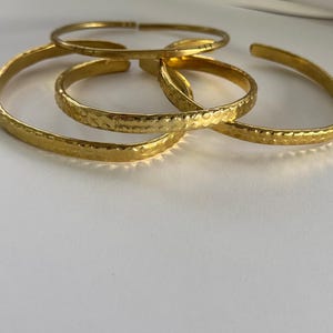 May include: Four gold-toned hammered metal bangles stacked together. The bangles are all the same width and have a slightly textured surface.