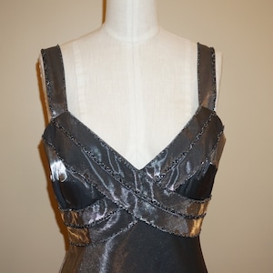 May include: A black sleeveless dress with a V-neckline and a criss-cross design on the bodice. The dress is made of a shiny fabric and has a beaded trim.