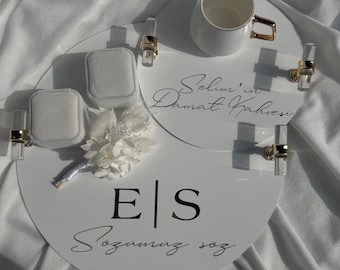 Puede incluir: Bandeja circular blanca con detalles dorados y dos pequeñas cajas blancas. La bandeja tiene el texto "Selim'in Damat Kahvesi" y "E|S Sozümüz söz" escrito en ella. Una taza de café blanca con asa dorada está sobre una bandeja circular blanca más pequeña.