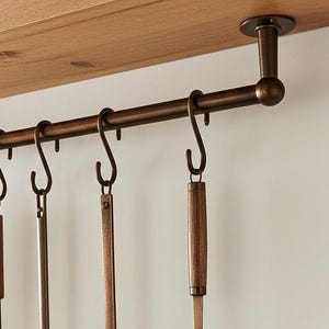 May include: A bronze kitchen utensil rack with hanging hooks, holding a white tea towel and four cooking tools. The tools include a slotted spoon, a spatula, and two ladles. The rack is mounted under a wooden shelf.