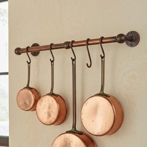 May include: A copper pot rack with four copper pans suspended from hooks. The pans vary in size and have long handles. The rack is fixed to a beige wall. A copper pan with a wooden spoon and a black spoon is on a wooden surface.