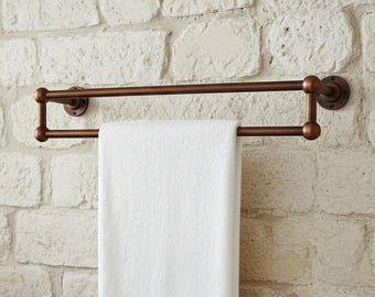 Puede incluir: Un toallero de color bronce montado en una pared de ladrillo texturizado de color blanquecino. Una toalla blanca doblada cuelga del toallero. Una bañera de color cobre es parcialmente visible en la esquina inferior izquierda.