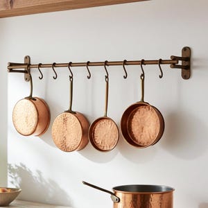 Könnte beinhalten: Kupfer-Kochgeschirr-Set, das an einer bronzefarbenen Stange an einer weißen Wand hängt. Das Set umfasst mehrere gehämmerten Kupferpfannen mit bronzefarbenen Griffen. Ein Kupfertopf steht auf einer Holz-Arbeitsplatte, neben einer Obstschale und einer Blumenvase.
