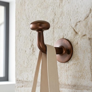 Könnte beinhalten: Ein bronzefarbener Wandhaken mit abgerundeter Oberseite und gebogenem Arm, montiert auf einer kreisförmigen Basis. Ein beigefarbener Riemen hängt an dem Haken. Der Haken ist an einer strukturierten, hellen Steinmauer befestigt.