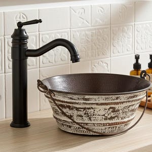 May include: A rustic bathroom sink with a hammered copper interior and distressed white exterior. The sink has a metal handle and is paired with a black faucet. The background features white tiles with floral patterns.