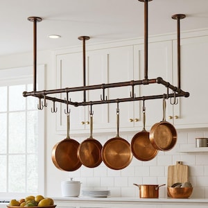 Może przedstawiać: Miedziany stojak na garnki zawieszony pod sufitem, trzymający kilka miedzianych patelni. Stojak wykonany jest z ciemnych metalowych rur i posiada wiele haczyków. Kuchnia posiada białe szafki i drewniany blat.