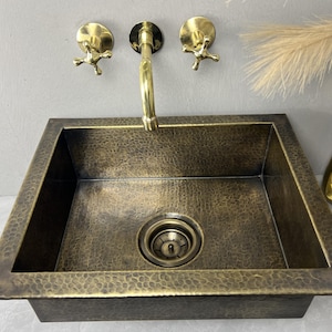 May include: A square, hammered copper sink with a bronze finish. The sink has a central drain and is paired with a gold faucet and matching wall-mounted fixtures. The design is rustic and elegant, suitable for a bathroom or kitchen.