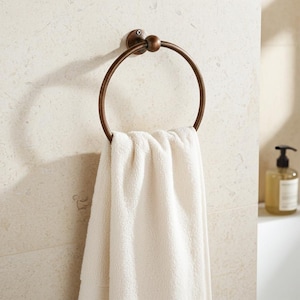 Könnte beinhalten: Ein bronzefarbener Handtuchring mit kreisförmigem Design, montiert an einer hellen Wand. Ein weißes Handtuch hängt an dem Ring. Eine Seifenflasche ist im Hintergrund zu sehen.