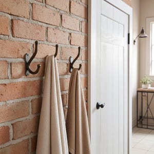 May include: Three bronze-coloured metal hooks mounted on a red brick wall, each holding a beige towel. A white door is visible on the right side of the image, with a black door handle. A small side table and a wall-mounted lamp are also in view.