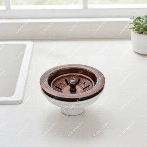 May include: A bronze-colored kitchen sink strainer with a white base. The strainer has a circular design with a central knob and a series of drainage holes. The product is designed to fit into a standard kitchen sink drain.