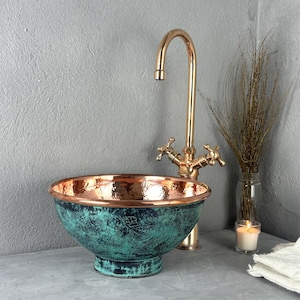 May include: A copper bathroom sink with a green patina finish and a matching copper faucet. The sink is round and has a wide, shallow bowl. The faucet has a single handle and a high spout.