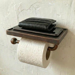 May include: A bronze-toned toilet paper holder with a shelf. A black wallet and a smartphone rest on the shelf. A roll of white, patterned toilet paper is in place. The holder is mounted on a textured wall.