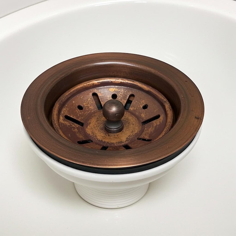 May include: A copper-toned kitchen sink drain assembly with a round strainer and a white ceramic base. The strainer has a central knob and multiple drainage holes. The assembly is set against a white background.