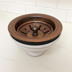 May include: A copper-toned kitchen sink strainer with a round, perforated design. The strainer has a dark rubber seal and sits atop a white, ceramic base. The overall design is functional and decorative, suitable for kitchen use.