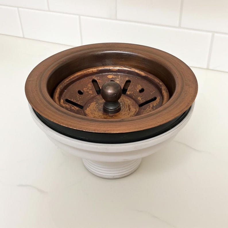 May include: A copper-toned kitchen sink strainer with a round, perforated design. The strainer has a dark rubber seal and sits atop a white, ceramic base. The overall design is functional and decorative, suitable for kitchen use.