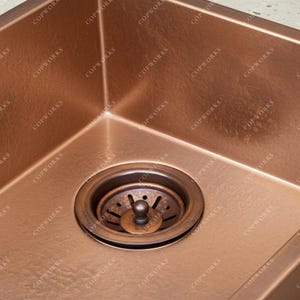 May include: A copper kitchen sink with a drain. The sink has a square shape with rounded corners. The drain is made of copper and has a circular shape with a strainer. The sink is a warm, reddish-brown color.