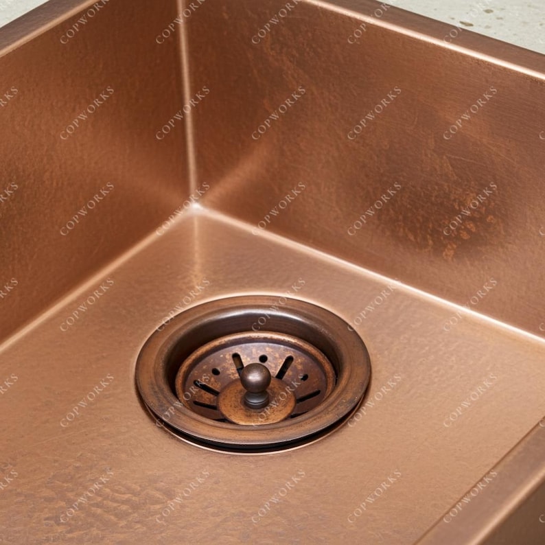 May include: A copper kitchen sink with a drain. The sink has a square shape with rounded corners. The drain is made of copper and has a circular shape with a strainer. The sink is a warm, reddish-brown color.