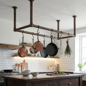 Puede incluir: Un estante para ollas suspendido del techo, hecho de tuberías de metal color cobre. Sostiene varias ollas y sartenes, incluyendo utensilios de cocina de cobre y hierro fundido negro. Un batidor y hierbas cuelgan del estante. La cocina tiene azulejos blancos.