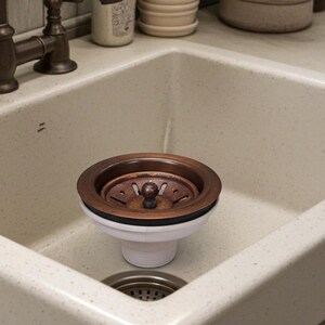 May include: A bronze-colored kitchen sink drain with a white base and a small, round, bronze-colored stopper. The drain is set in a speckled, off-white sink.