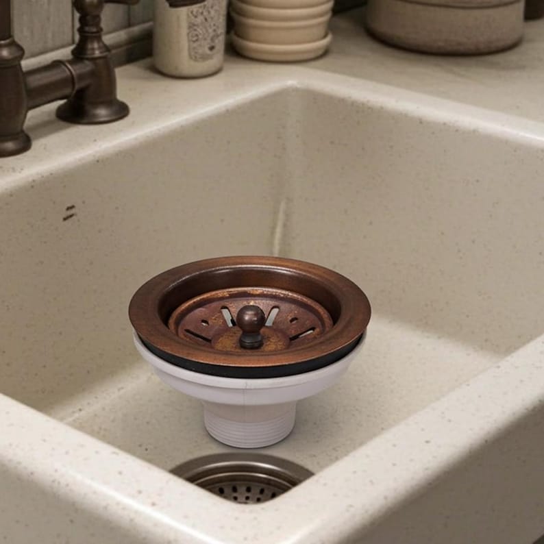 May include: A bronze-colored kitchen sink drain with a white base and a small, round, bronze-colored stopper. The drain is set in a speckled, off-white sink.