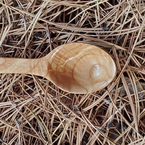 May include: A wooden spoon with a natural finish, lying on a bed of brown pine needles.