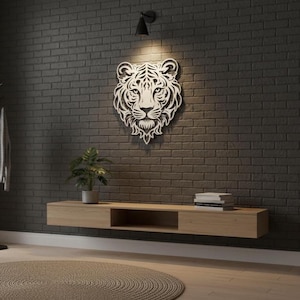May include: A white, laser-cut tiger head wall art piece on a dark grey brick wall. Below, a light wood floating shelf holds a potted plant and books. A black spotlight shines on the tiger.