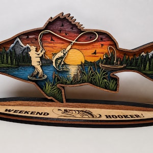 Puede incluir: Una obra de arte de madera en capas con forma de pez, que representa una escena de pesca. La obra presenta un pescador, un pez saltando fuera del agua, un barco y una puesta de sol. La base dice "WEEKEND HOOKERS".