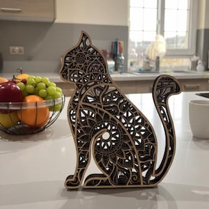 Puede incluir: Silueta decorativa de gato de madera con intrincados cortes estilo mandala. El gato es marrón con detalles negros, sentado erguido con la cola enroscada. Un artículo de decoración del hogar único.