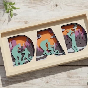 Puede incluir: Obra de arte enmarcada en madera con la palabra "DAD", con recortes de papel en capas de un padre y un niño en varias actividades. El fondo tiene tonos naranja, morado y gris. Incluye una pequeña etiqueta "Handmade by Studio A".