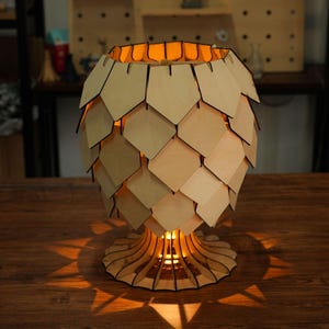Puede incluir: Una lámpara de mesa construida en madera de color claro, con una pantalla compuesta por escamas pentagonales superpuestas. La base es circular con listones radiantes y el interior emite una luz cálida que proyecta sombras.