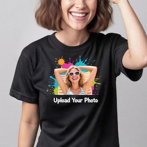 Puede incluir: Una camiseta negra de manga corta con un estampado gráfico central. El diseño muestra un fondo de salpicaduras de pintura coloridas con la imagen fotográfica de una persona sonriente con gafas de sol, sobre el texto blanco Upload Your Photo.