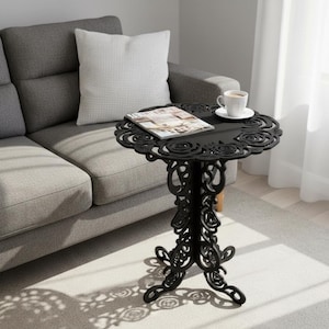 Puede incluir: Mesa auxiliar negra y ornamentada con un diseño floral de rosas. La mesa tiene una parte superior redonda y una base decorativa. Una revista y una taza de café están sobre la mesa, junto a un sofá gris.